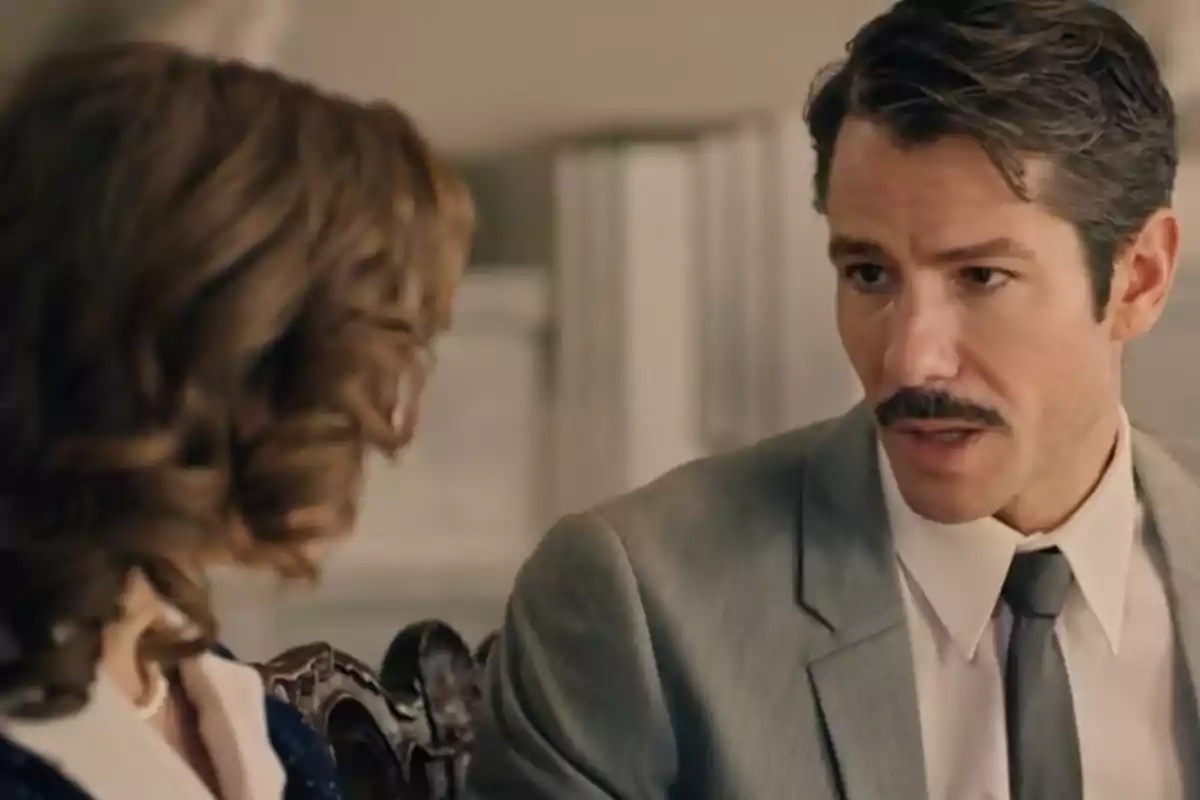 Pelai amb bigoti i vestit gris parlant amb una dona de cabells arrissats en un ambient interior elegant. Captura de 'Sueños de libertad' Pelai amb bigoti i vestit gris parlant amb una dona de cabells arrissats en un ambient interior elegant. Captura de 'Sueños de libertad'