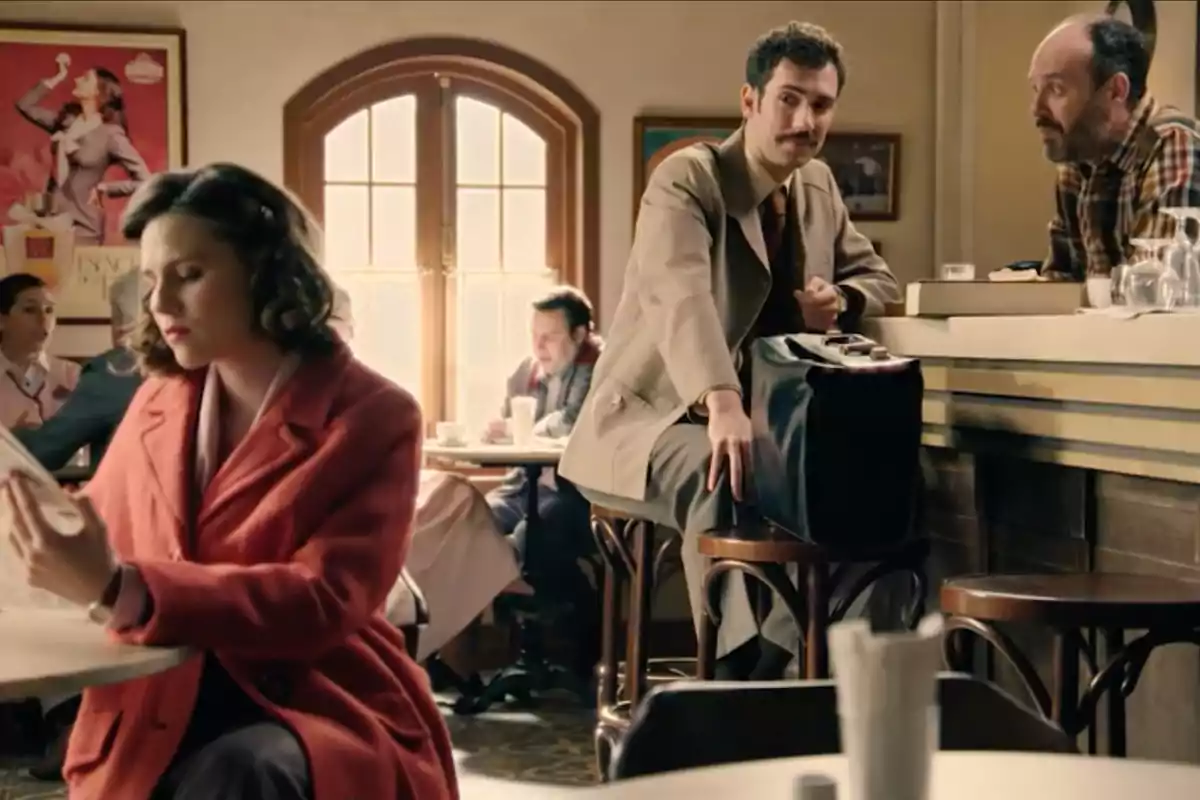 Diverses persones són en una cafeteria d’estil antic, una dona amb abric vermell llegeix un diari en primer pla mentre un home amb bigoti i vestit clar seu a la barra conversant amb el cambrer. Captura de ‘somnis de llibertat’ Diverses persones són en una cafeteria d’estil antic, una dona amb abric vermell llegeix un diari en primer pla mentre un home amb bigoti i vestit clar seu a la barra conversant amb el cambrer. Captura de ‘somnis de llibertat’