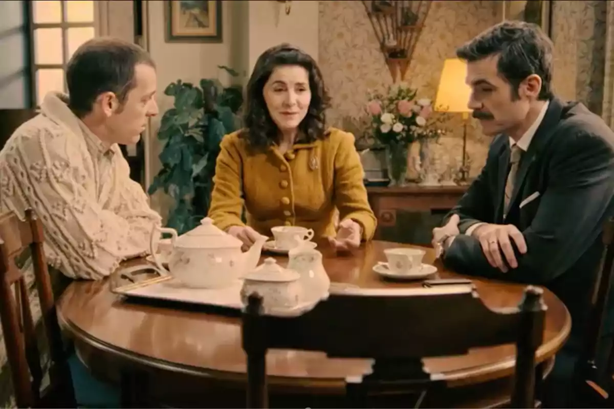 Luis, Digna i Joaquín asseguts al voltant d’una taula rodona de fusta prenent te en una sala decorada amb flors i una làmpada encesa al fons. Captura de ‘Sueños de libertad’ Luis, Digna i Joaquín asseguts al voltant d’una taula rodona de fusta prenent te en una sala decorada amb flors i una làmpada encesa al fons. Captura de ‘Sueños de libertad’