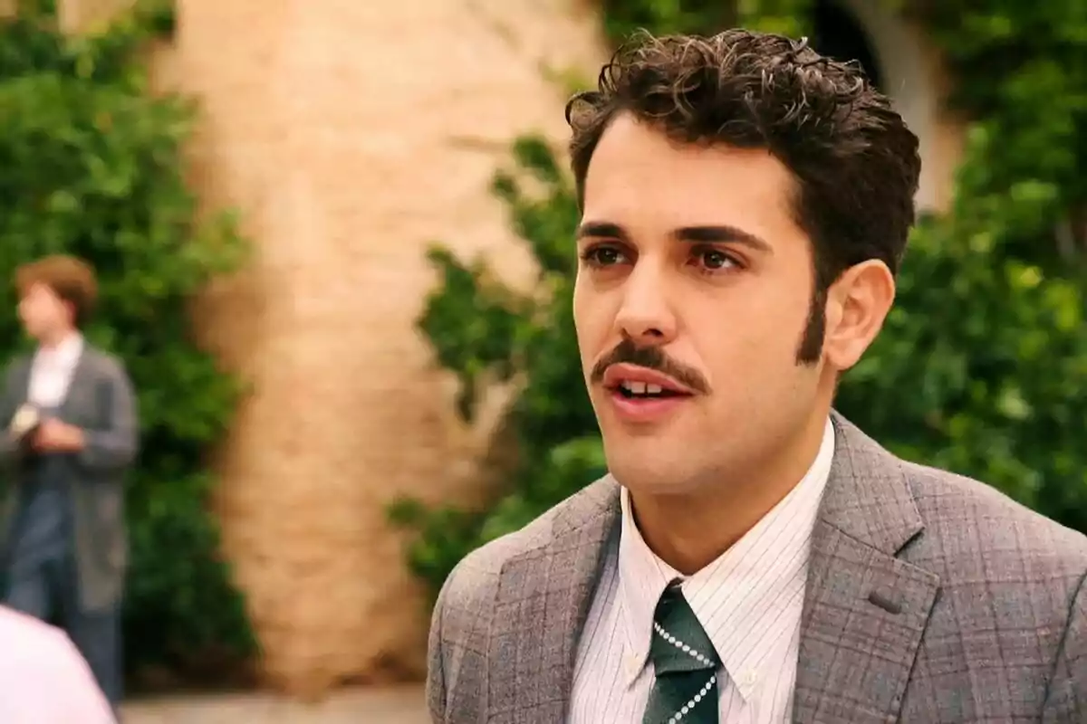 Santiago con bigote y traje a cuadros está al aire libre con un fondo de vegetación y una pared de ladrillo. Captura de 'Sueños de libertad'