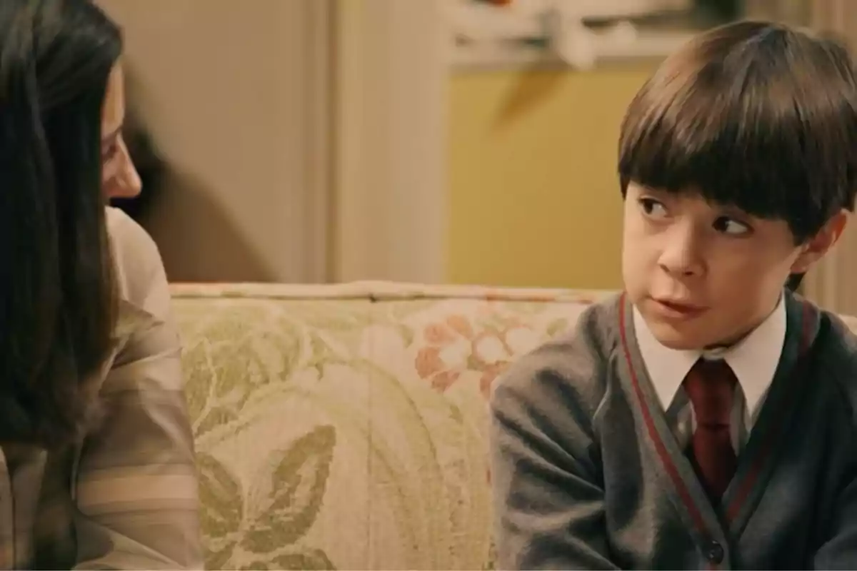 Teo amb uniforme escolar i corbata vermella està assegut en un sofà estampat al costat d’una dona, tots dos mirant-se i conversant en una sala il·luminada. Captura de ‘Sueños de libertad’ Teo amb uniforme escolar i corbata vermella està assegut en un sofà estampat al costat d’una dona, tots dos mirant-se i conversant en una sala il·luminada. Captura de ‘Sueños de libertad’
