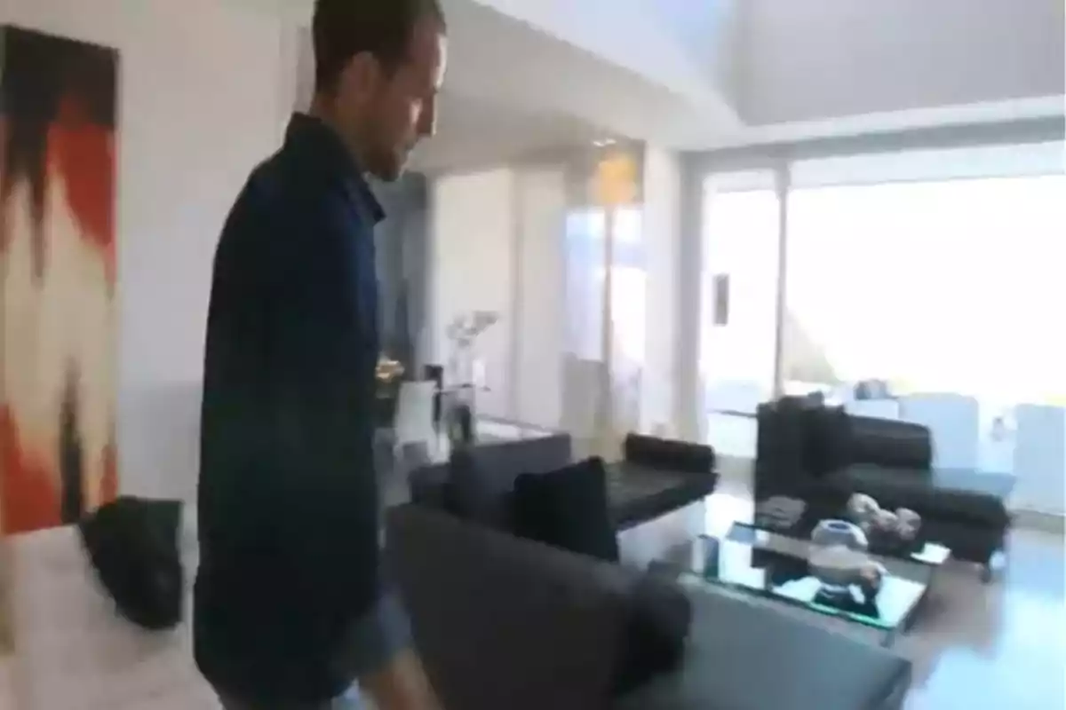 Un home camina per una sala d'estar moderna amb sofàs foscos i una gran finestra. Un home camina per una sala d'estar moderna amb sofàs foscos i una gran finestra.