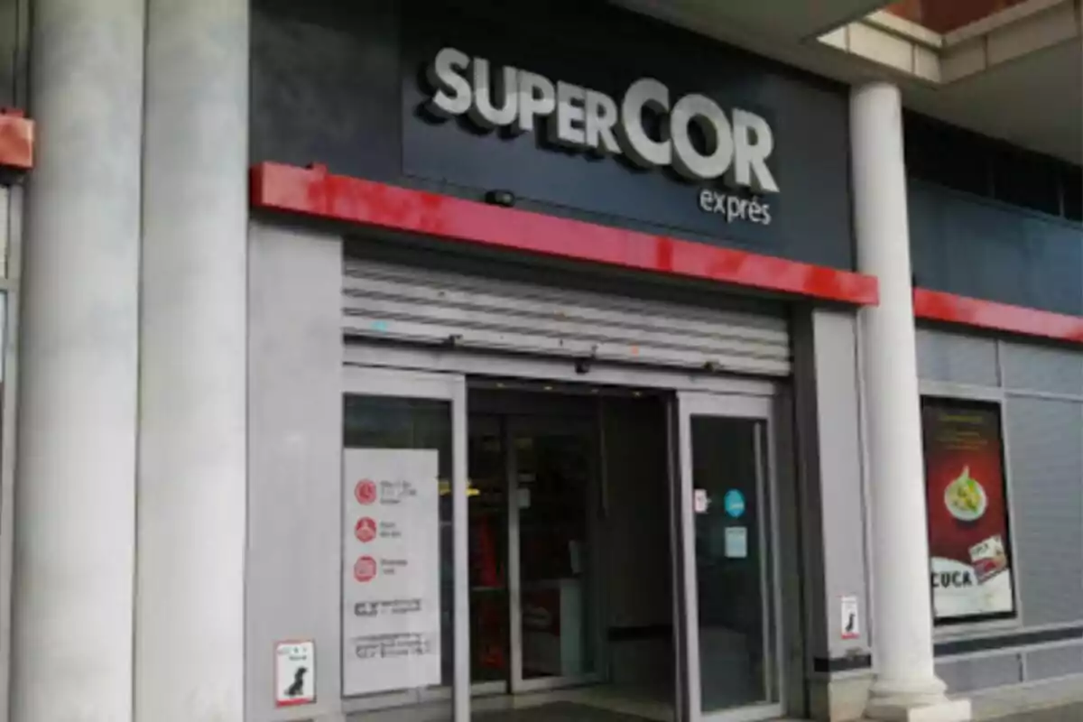 Entrada d´una botiga Supercor Exprés amb portes de vidre i cartells a la façana. Entrada d´una botiga Supercor Exprés amb portes de vidre i cartells a la façana.