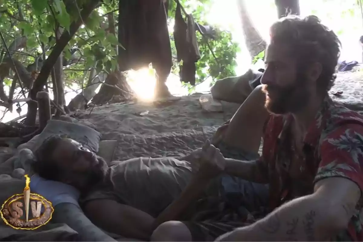 Álvaro Muñoz Escassi i Pelayo Díaz es donen la mà en una platja sota l’ombra dels arbres, un està estirat i l’altre assegut, amb llum de sol filtrant-se entre les fulles i roba penjada al fons. Captura de 'Supervivientes' Álvaro Muñoz Escassi i Pelayo Díaz es donen la mà en una platja sota l’ombra dels arbres, un està estirat i l’altre assegut, amb llum de sol filtrant-se entre les fulles i roba penjada al fons. Captura de 'Supervivientes'