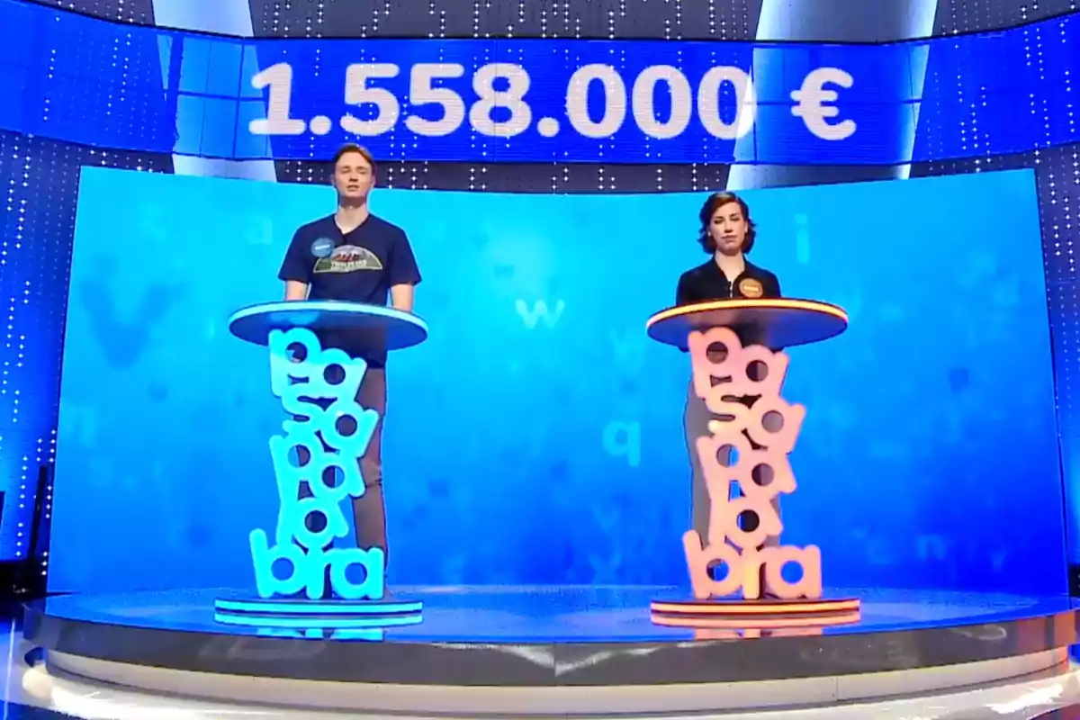 Manu i Rosa estan drets en un plató de televisió amb un fons blau, cadascun darrere d'un faristol decorat amb lletres, i a la part superior es mostra un marcador amb una quantitat en euros. Captura de 'Pasapalabra'. Manu i Rosa estan drets en un plató de televisió amb un fons blau, cadascun darrere d'un faristol decorat amb lletres, i a la part superior es mostra un marcador amb una quantitat en euros. Captura de 'Pasapalabra'.