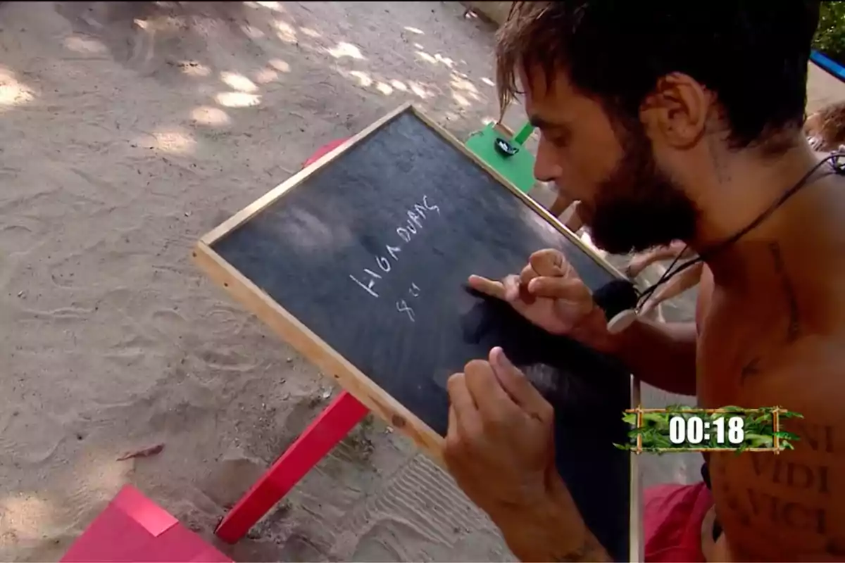 Montoya escriu en una pissarra petita mentre està assegut a la sorra amb un cronòmetre a la cantonada inferior dreta mostrant 00:18. Captura de 'Supervivientes' Montoya escriu en una pissarra petita mentre està assegut a la sorra amb un cronòmetre a la cantonada inferior dreta mostrant 00:18. Captura de 'Supervivientes'