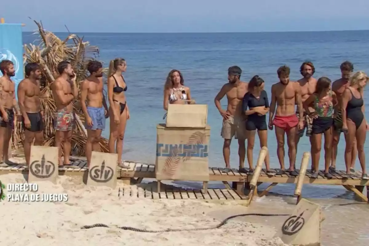 Un grup de persones en banyador està reunit en una platja al costat d'un podi de fusta, amb el mar de fons i un cartell blau a l'esquerra. Captura de 'Supervivientes' Un grup de persones en banyador està reunit en una platja al costat d'un podi de fusta, amb el mar de fons i un cartell blau a l'esquerra. Captura de 'Supervivientes'