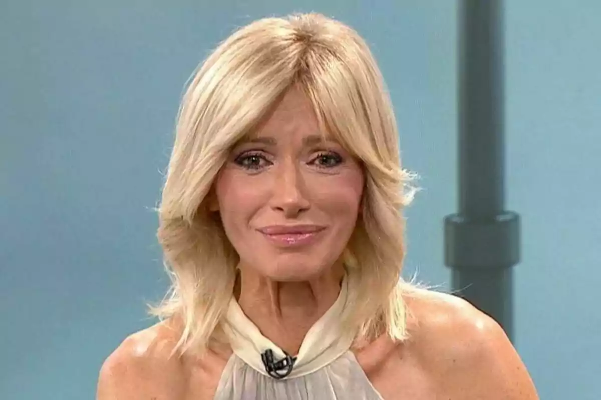 Captura de Susanna Griso rubia con cabello corto y ondulado, usando un vestido sin mangas y un micrófono de solapa, con una expresión facial seria y un fondo azul claro en Espejo Público