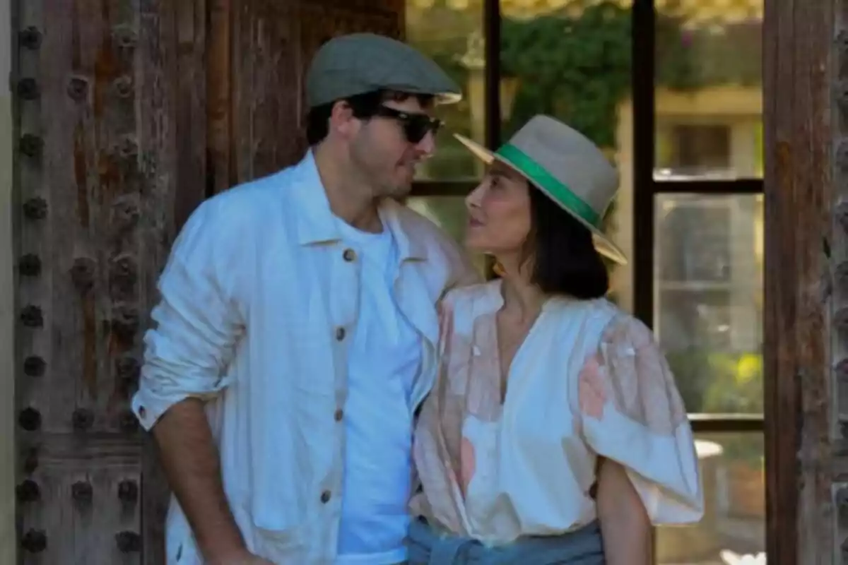 Una pareja -formada por Íñigo Onieva y Tamara Falcó- mirándose con cariño mientras posan juntos al aire libre frente a una puerta de madera.