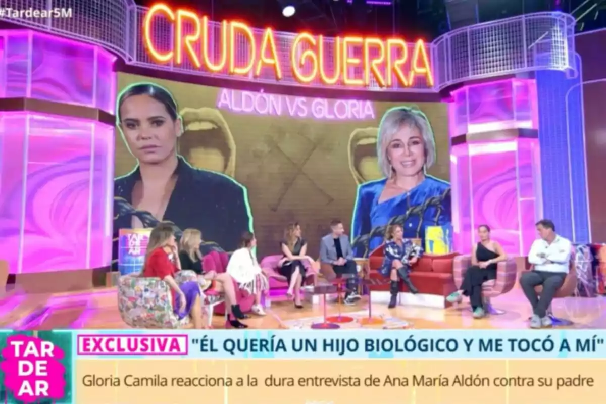 Un grup de persones està assegut en un plató de televisió acolorit, amb un gran cartell que diu Un grup de persones està assegut en un plató de televisió acolorit, amb un gran cartell que diu
