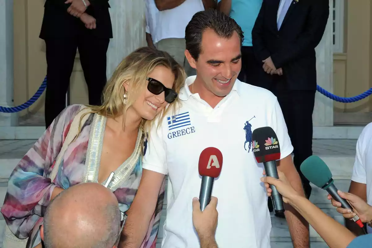 Tatiana Blatnik i Nicolau de Grècia somrient és entrevistada per diversos periodistes amb micròfons en un esdeveniment a l'aire lliure. Tatiana Blatnik i Nicolau de Grècia somrient és entrevistada per diversos periodistes amb micròfons en un esdeveniment a l'aire lliure.