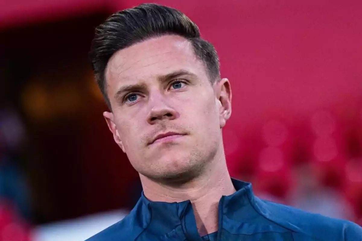 A man with short dark hair, wearing a blue jacket, with a serious expression and a red blurred background.