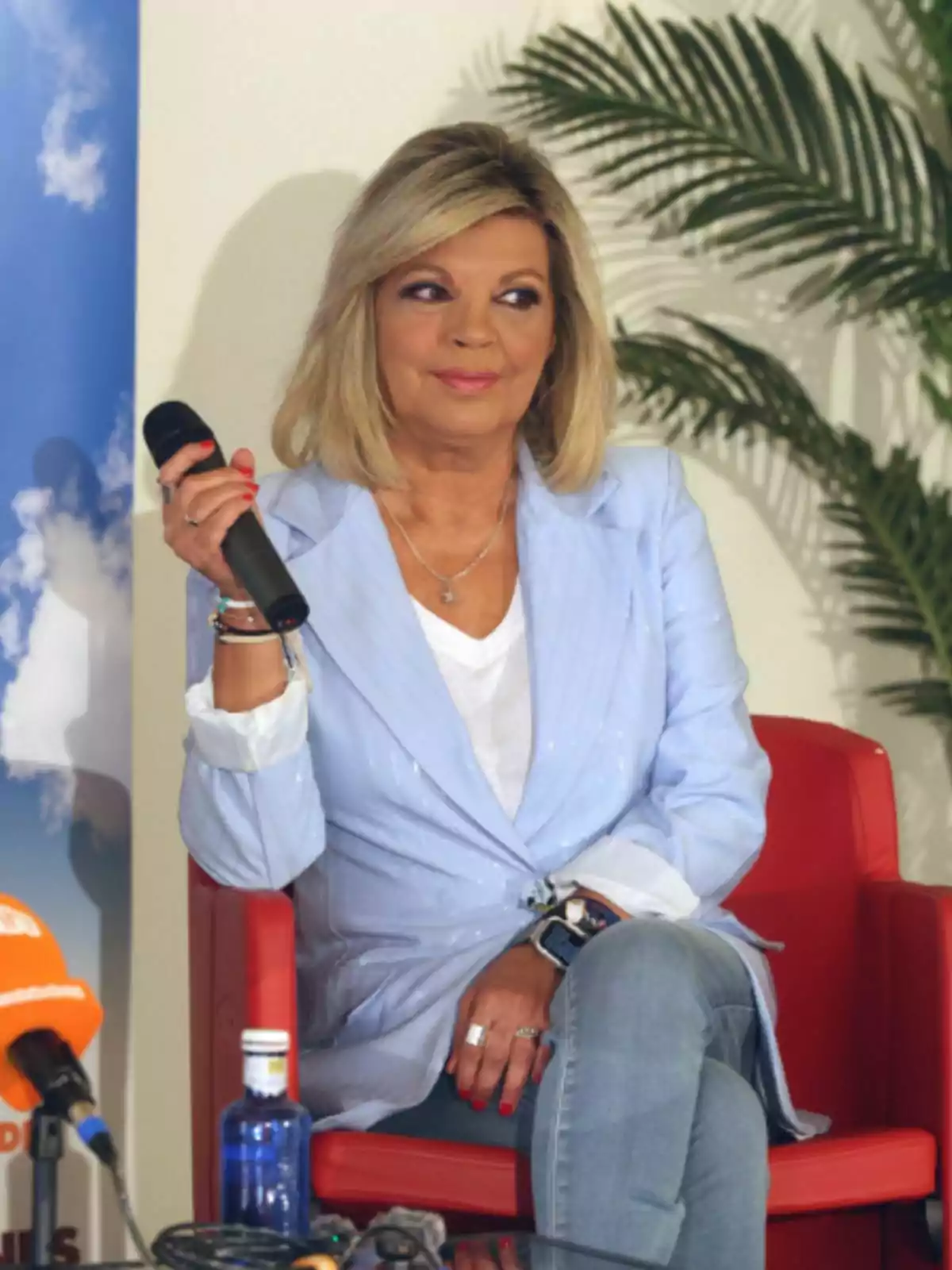 Terelu Campos sitting on a red chair holding a microphone, wearing a light blue jacket and denim pants, with a background of plants and microphones on the table. Terelu Campos sitting on a red chair holding a microphone, wearing a light blue jacket and denim pants, with a background of plants and microphones on the table.