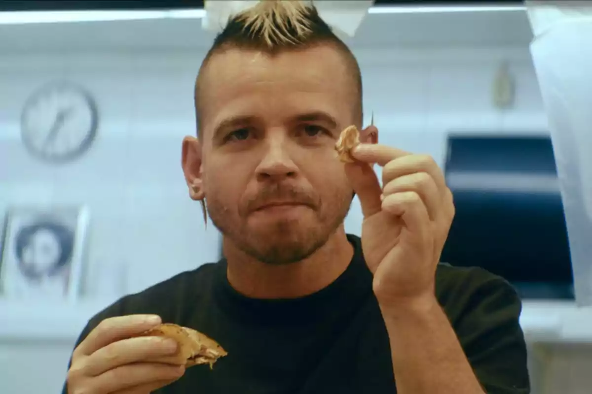 Dabiz Muñoz amb pentinat modern sostenint menjar en una mà i un tros petit a l'altra en un entorn de cuina a Univerxo Dabiz. Dabiz Muñoz amb pentinat modern sostenint menjar en una mà i un tros petit a l'altra en un entorn de cuina a Univerxo Dabiz.