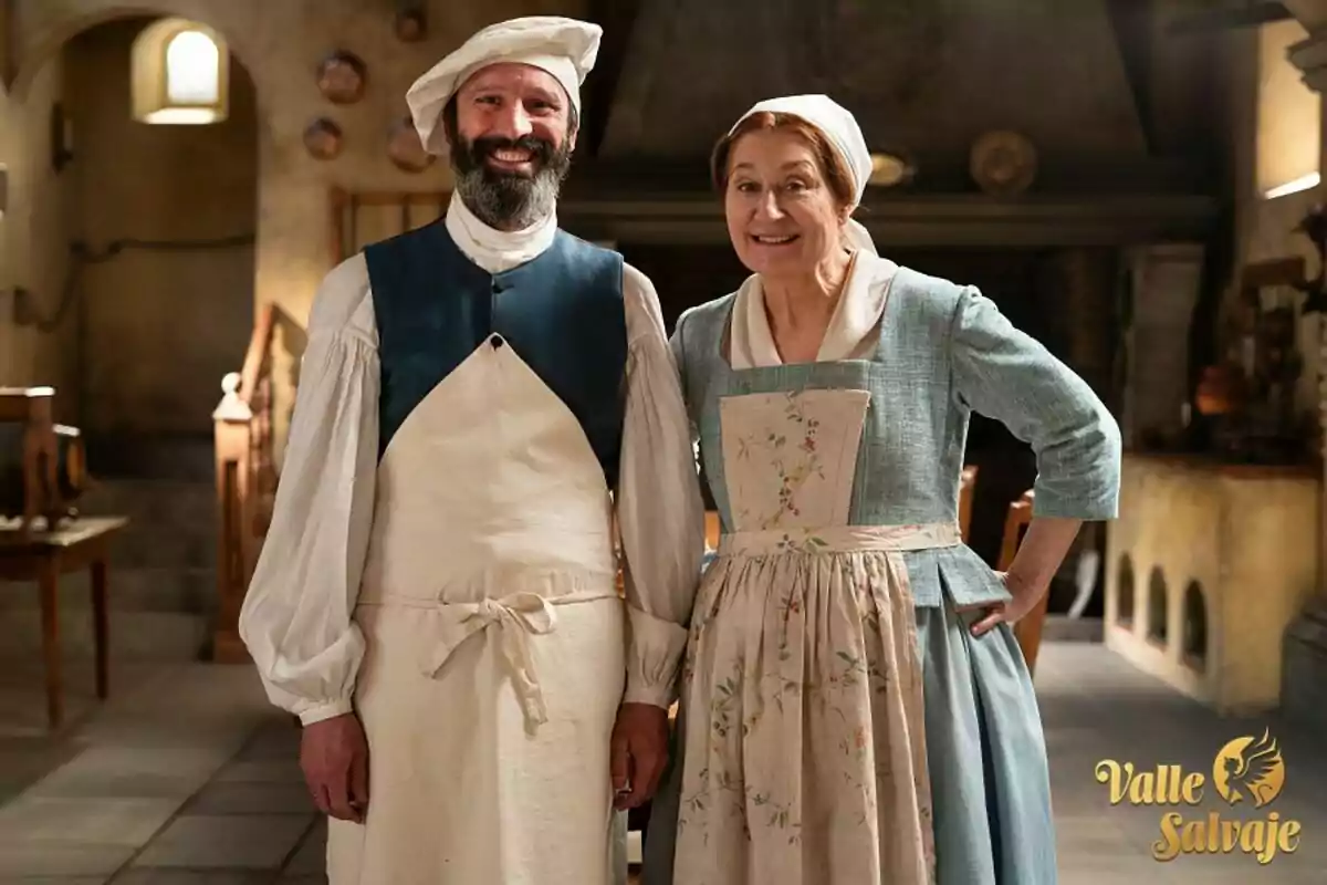 Amadeu i Eva vestida amb roba d'època, tots dos somrients, a l'interior d'una cuina antiga amb utensilis de cuina al fons. Amadeu i Eva vestida amb roba d'època, tots dos somrients, a l'interior d'una cuina antiga amb utensilis de cuina al fons.