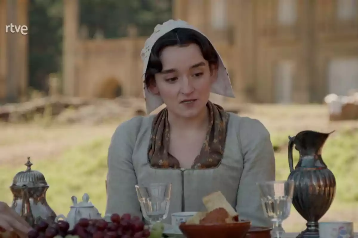 Matilde vestida con ropa de época y un pañuelo en la cabeza está sentada en una mesa al aire libre con comida y bebida, en un entorno campestre con un edificio antiguo de fondo. Captura de 'Valle Salvaje'