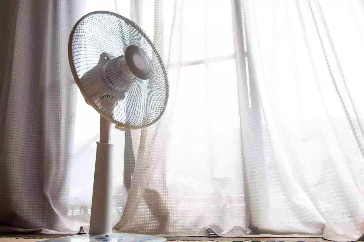 Un ventilador davant d'una finestra amb cortina blanca