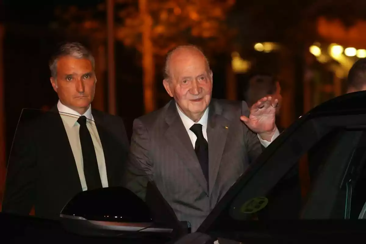 Juan Carlos I en traje gris saluda mientras está de pie junto a un automóvil, acompañado por Vicente García-Mochales en traje oscuro.