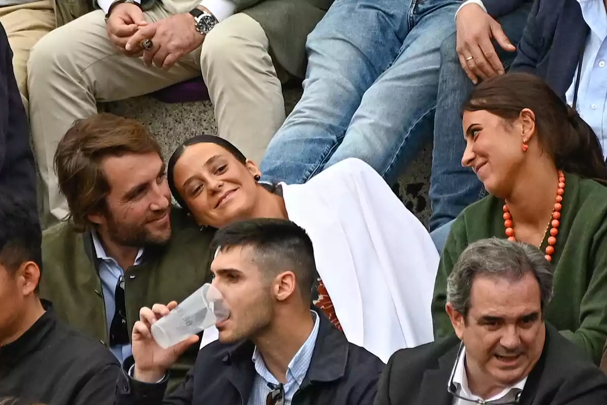 Un grup de persones assegudes a les grades, algunes conversant i somrient, mentre una persona beu d'un got. Un grup de persones assegudes a les grades, algunes conversant i somrient, mentre una persona beu d'un got.