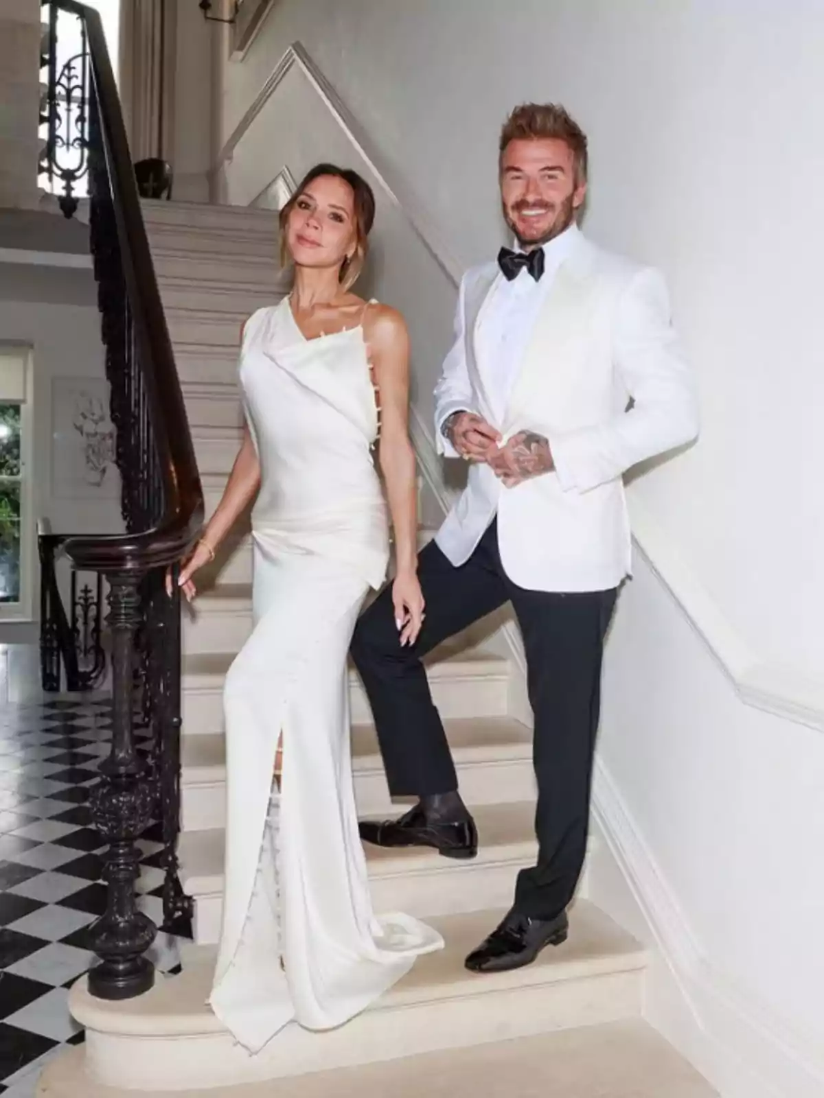 Una parella elegantament vestida posa en una escala dins d’una casa, tots dos porten roba formal en tons blancs i negres. Una parella elegantament vestida posa en una escala dins d’una casa, tots dos porten roba formal en tons blancs i negres.