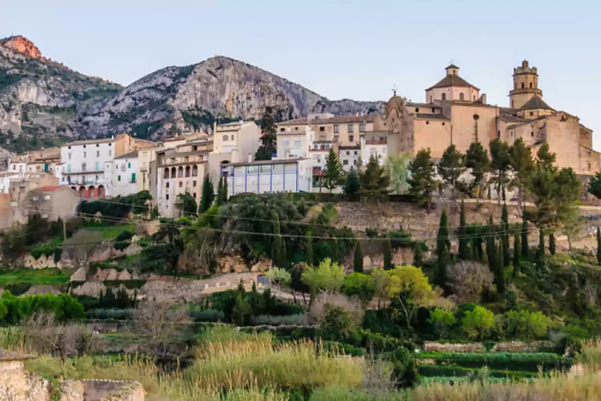 Vista panoràmica d´un poble amb una església en un turó envoltat de muntanyes i vegetació.