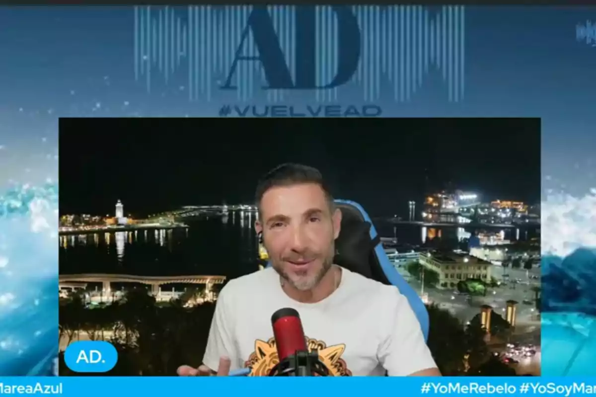 Antonio David Flores amb una samarreta clara parlant davant d’un micròfon vermell amb un fons nocturn d’una ciutat il·luminada i el mar Antonio David Flores amb una samarreta clara parlant davant d’un micròfon vermell amb un fons nocturn d’una ciutat il·luminada i el mar