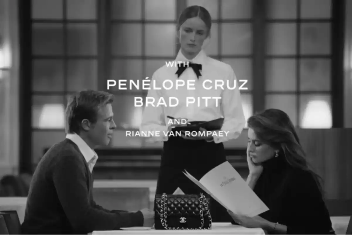 A black and white scene shows Brad Pitt and Penélope Cruz sitting at a restaurant table, a woman is reading a menu while a man watches her, a waitress standing holds a notepad, in the center of the table there is a designer handbag. A black and white scene shows Brad Pitt and Penélope Cruz sitting at a restaurant table, a woman is reading a menu while a man watches her, a waitress standing holds a notepad, in the center of the table there is a designer handbag.