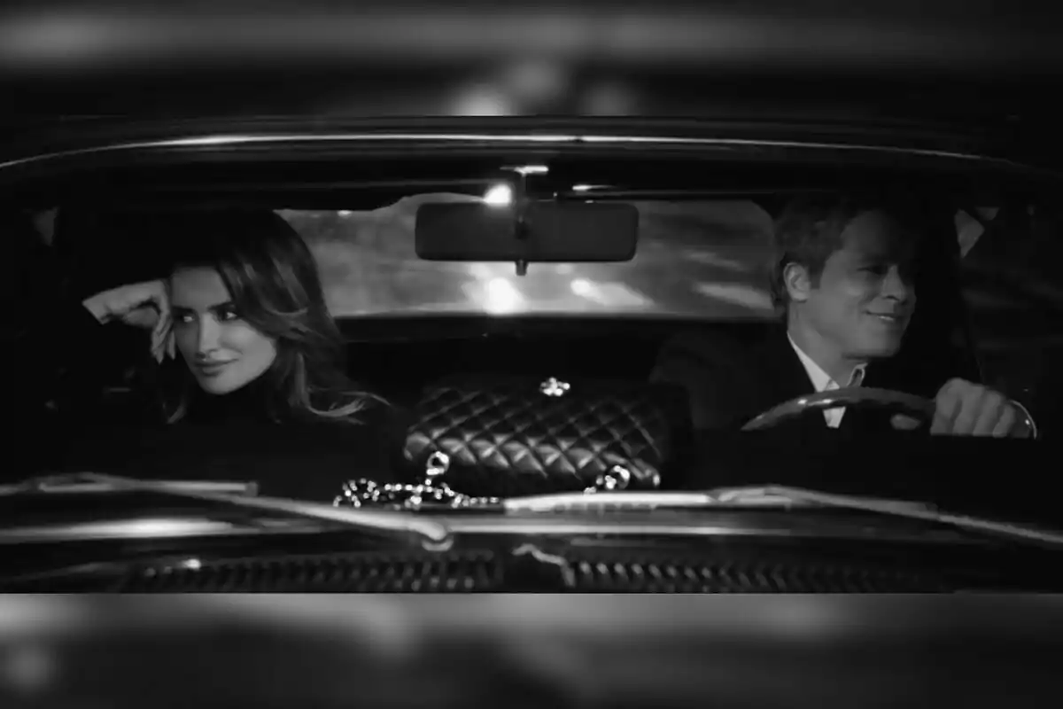 Penélope Cruz and Brad Pitt smile while sitting in a classic car in a black and white image. Penélope Cruz and Brad Pitt smile while sitting in a classic car in a black and white image.