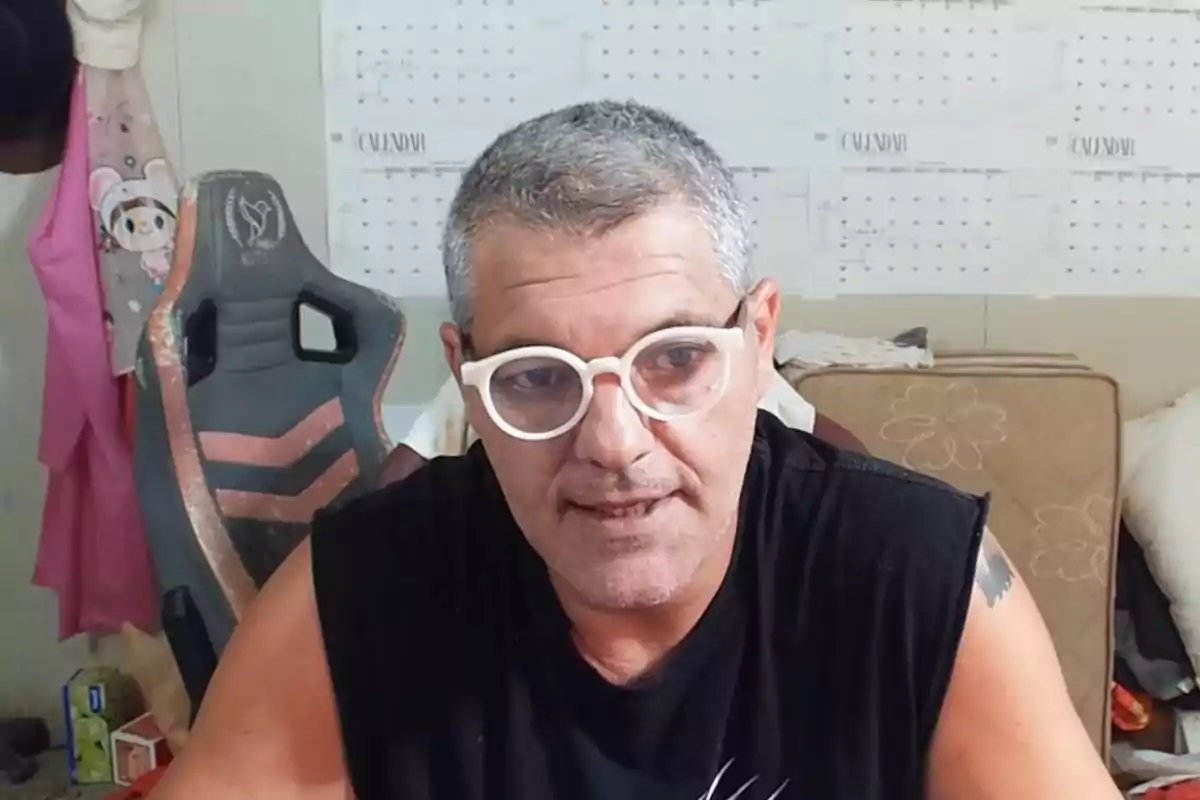 Frank Blanco amb ulleres blanques i samarreta negra està assegut davant d’un calendari a la paret, amb una cadira de joc al seu costat. Frank Blanco amb ulleres blanques i samarreta negra està assegut davant d’un calendari a la paret, amb una cadira de joc al seu costat.