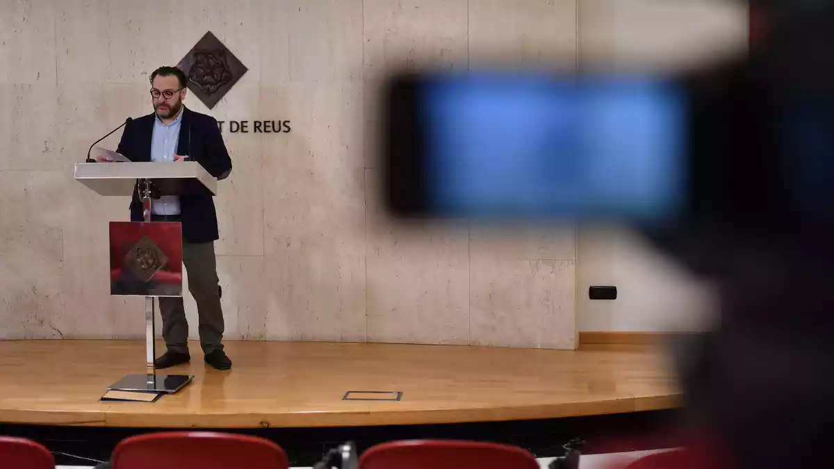 Daniel Recasens, al fons, a la sala de premsa, amb una càmera de vídeo borrosa en primer pla