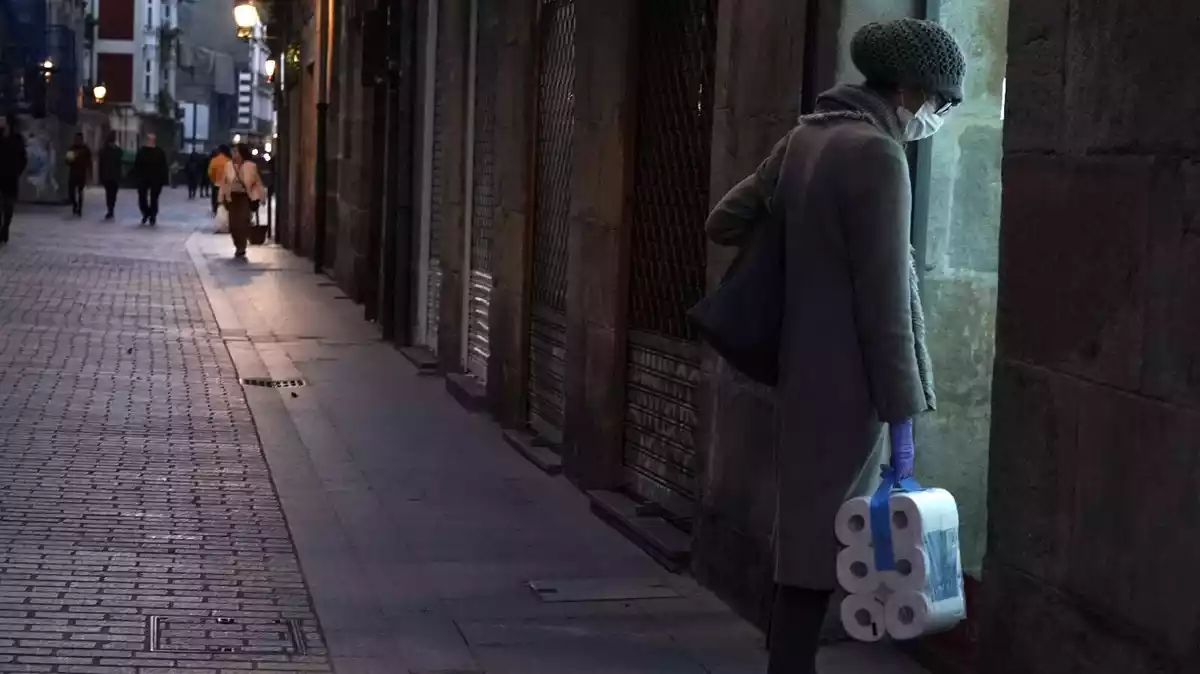 Una dona, protegida amb una mascareta, transporta un paquet de paper de vàter en un carrer fosc