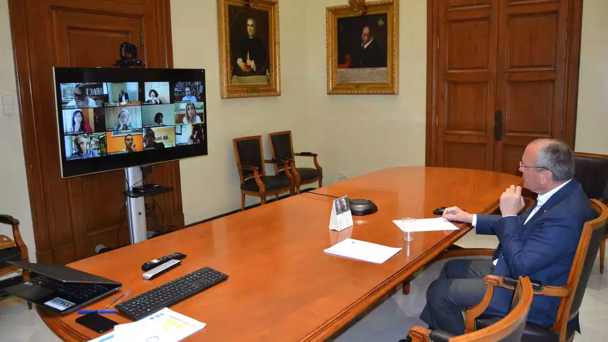 Carles Pellicer assegut a l'Ajuntament amb una pantalla amb els participants a la reunió