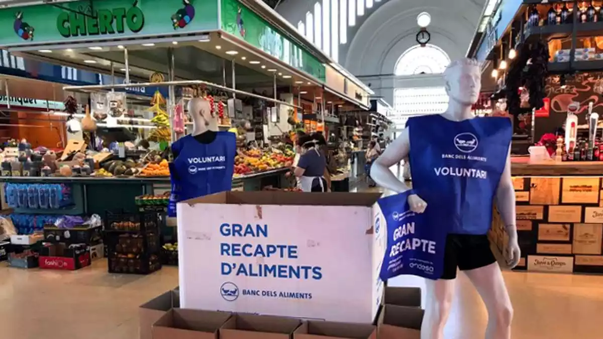 Els habituals dipòsits per a la recollida del Banc dels Aliments al mercat de Tortosa, amb un maniquí que substitueix els voluntaris, que no poden estar presents per la pandèmia